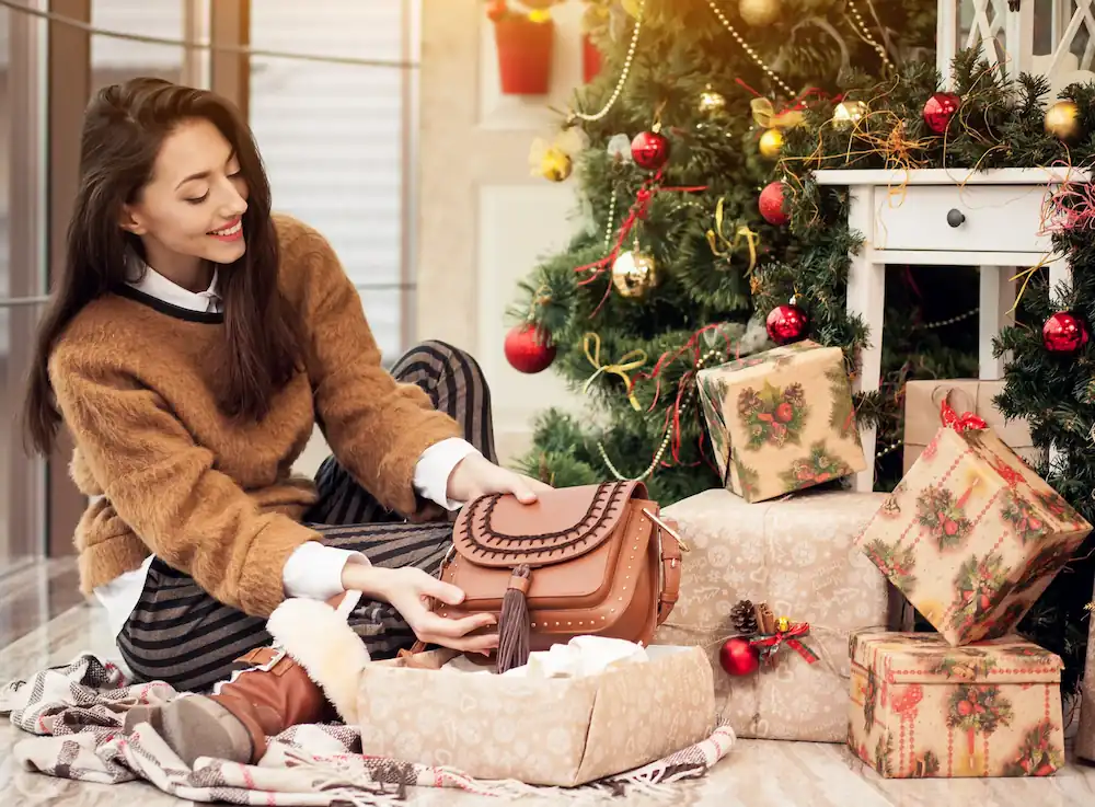 Woman unwrapping gifts under a decorated Christmas tree, symbolizing thoughtful gifting ideas with Gaia in Love.