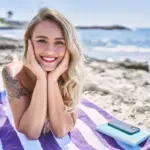 A cheerful young blonde woman lying on a striped beach towel with a radiant smile, enjoying the sunny outdoors. A perfect visual for Gaia in Love's guide on selling rabbit vobrator products on eBay Australia.