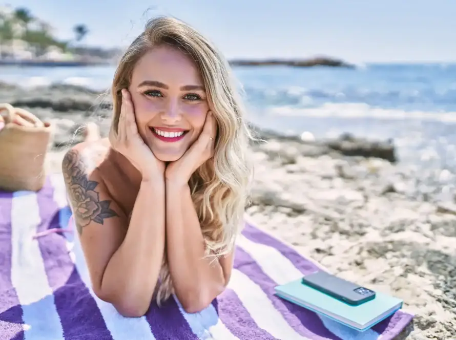 A cheerful young blonde woman lying on a striped beach towel with a radiant smile, enjoying the sunny outdoors. A perfect visual for Gaia in Love's guide on selling rabbit vibratoe products on eBay Australia.