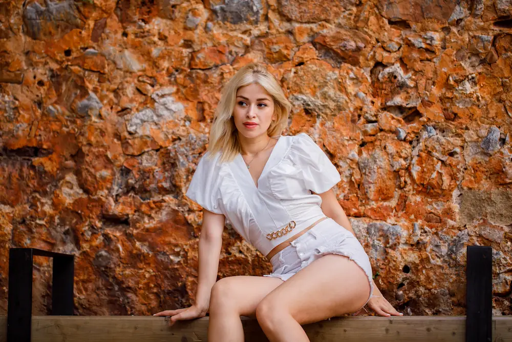 A blonde woman sitting by a rustic stone wall, representing the empowerment and inclusivity of Gaia in Love's best couples toys.