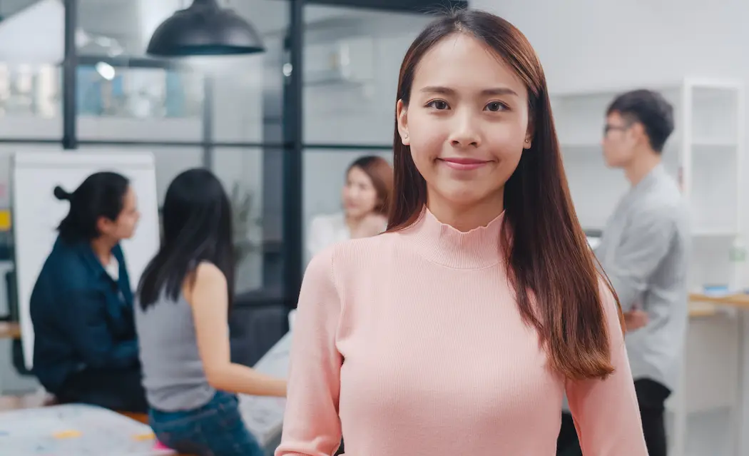 A confident young woman in a pink top smiles at the camera in an office setting, surrounded by colleagues engaged in lively discussions, reflecting Gaia in Love's inclusive and collaborative workplace culture.