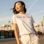 An Asian woman wearing a white T-shirt with "Gaia in Love" in violet, standing at a shipping port under soft sunlight, symbolizing China-US tariff agreement and sex toy export recovery.