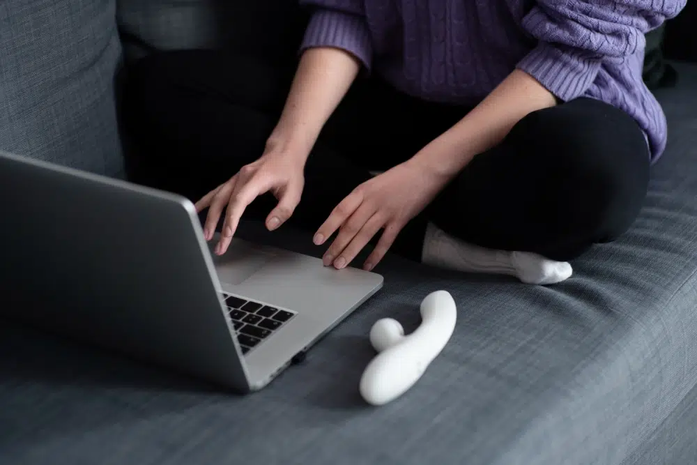 Person using a laptop on a couch with a white vibrator nearby, illustrating a better sex toy battery and charging experience.