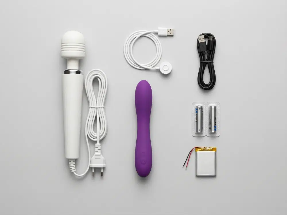 Flat lay of a plug-in wand massager, a rechargeable purple vibrator, USB and magnetic charging cables, AA batteries, and a Li-polymer battery cell on a gray background.